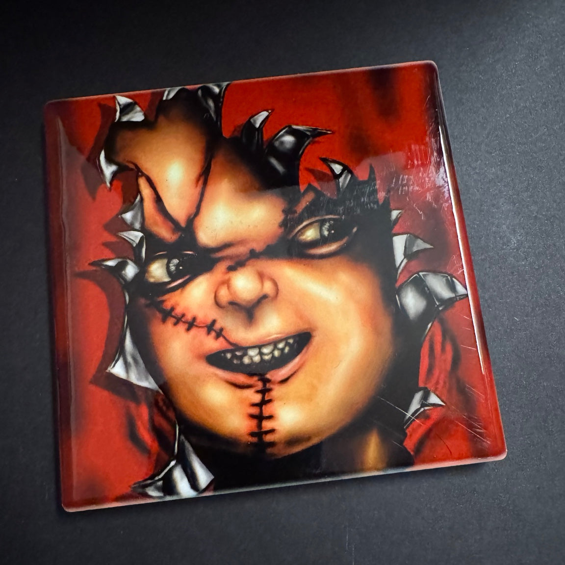 Square ceramic horror coaster with a sinister stitched doll face breaking through a vivid red background